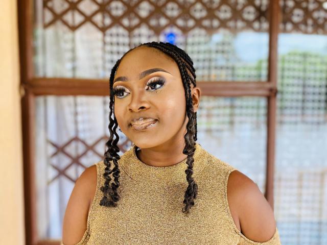 StaceyNoelle, a Black woman with dark brown skin, stands in front of a wooden-framed window with a lattice design. She has long, twisted braids falling to her shoulders. She wears a sleeveless, beige, textured top with cut-out shoulders. Her makeup includes dramatic, dark blue eyeliner and thick, black eyelashes. She has a subtle smile, and her lips are slightly parted. The background is slightly blurred, showing a mix of green and blue. Her expression is confident and relaxed.
