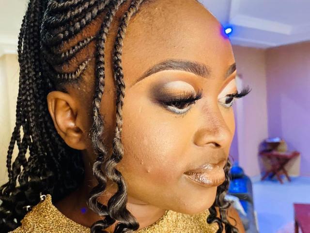 Close-up of StaceyNoelle with dark brown skin, braided black hair, and gold glitter top. StaceyNoelle has heavy, dramatic eye makeup with thick black eyeliner, white eyeshadow, and long, thick, black eyelashes. Her lips are glossy with a metallic sheen. The braids are neatly styled with a mix of straight and curly strands. The lighting highlights her smooth skin and glossy lips. The background is blurred, focusing on StaceyNoelle's face. The image is brightly lit with a blue light source in the upper right corner.