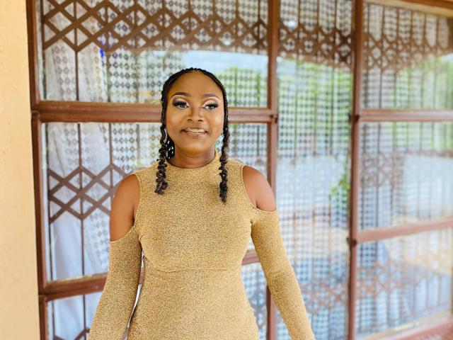 StaceyNoelle stands in front of a patterned wooden window. She has dark brown skin and wears a glittery, beige, off-shoulder dress with cutout sleeves. Her hair is in two neat braids, and she has glossy, dark brown eyes with winged eyeliner. She smiles slightly. The window has a geometric pattern with small, diamond-shaped designs. The photograph is bright with natural light. StaceyNoelle's makeup is bold and stylish. Her confident, casual pose adds to the image's modern, stylish vibe.