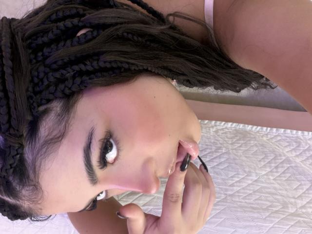 RheaVolk with braided hair, pink blush, and dark eyeliner, biting black nail with finger, pink bra visible, white quilted surface, looking up with big eyes, nose piercing.