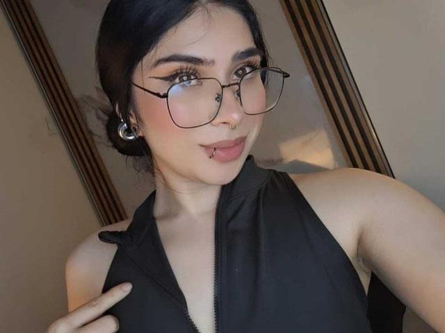 EmmaCollinsHot with glasses, black hair in a bun, nose ring, lip piercing, black sleeveless top, pointing at chest, fair skin, looking at camera, medium-sized tits, smiling.