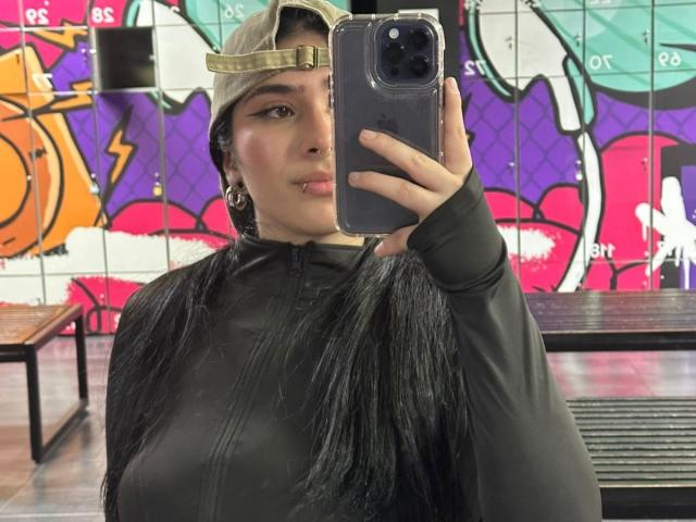 EmmaCollinsHot taking a selfie with a gold fronted baseball cap, black long-sleeve top, and black hair, holding a blue phone, showing her face and small tits.