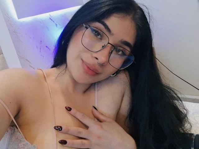 EmmaCollinsHot with long black hair, glasses, nose ring, and pink spaghetti straps, touching her tits with dark red, sparkly nails, smiling softly.