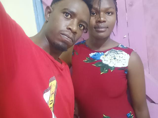 A photograph of two LykyBlacks standing close. The left LykyBlack has dark skin, short hair, and a goatee, wearing a red shirt with a graphic. The right LykyBlack has dark skin, braided hair, and wears a red floral dress with white and blue flowers. They both have neutral expressions and are slightly angled towards the camera. The background is a pale pink wall. The image is a casual selfie.