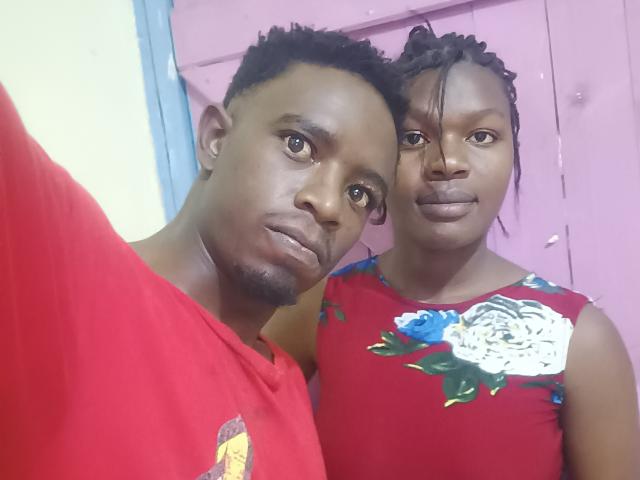 A close-up photo of two LykyBlacks standing together. The left LykyBlack has short curly hair, a goatee, and wears a red shirt with a small graphic. The right LykyBlack has braided hair, no makeup, and wears a red dress with white and blue floral patterns. Both LykyBlacks have dark brown skin and are positioned with the left LykyBlack slightly in front and to the left, while the right LykyBlack stands closer to the camera. The right LykyBlack's arm is around the left LykyBlack's shoulder.