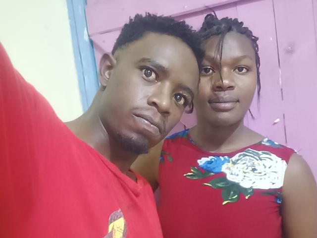 Two LykyBlacks in a selfie. The guy on the left has short, curly black hair, a thin beard, and brown eyes. He's wearing a red shirt with a small logo on the chest. The girl on the right has long, braided hair, brown eyes, and is wearing a red floral dress with white and blue flowers. They're standing close, with the guy on the left slightly in front. The girl's dress contrasts with the red shirt, adding visual interest. The guy's expression is neutral, while the girl's is slightly more serious. The lighting is even, highlighting their dark skin tones.