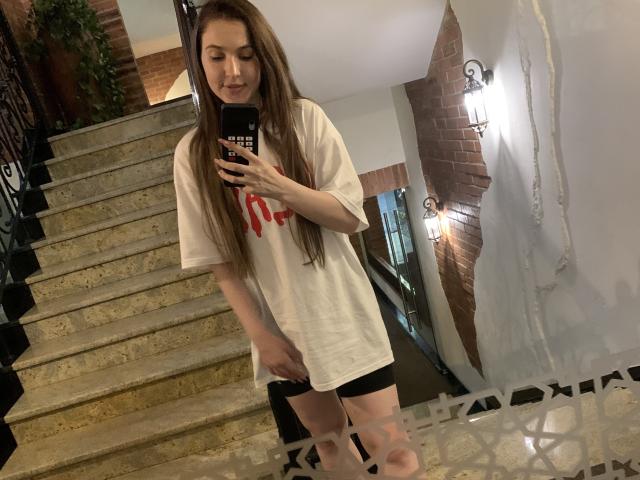 AliceAndCristal with long brown hair, wearing a white t-shirt and black shorts, standing on stairs, holding a phone, showing her pussy through the shorts. Mirror selfie.