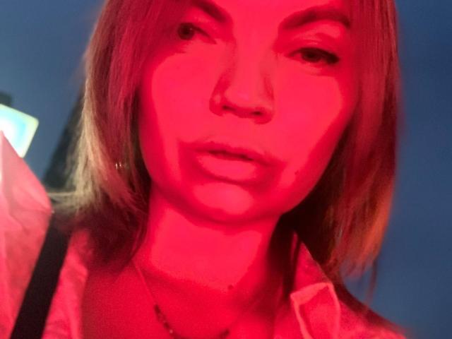 Red-lit selfie of ElizavetaCream with blonde hair, nose ring, and slightly parted lips, wearing a shirt with a necklace, and a strap visible on her shoulder.