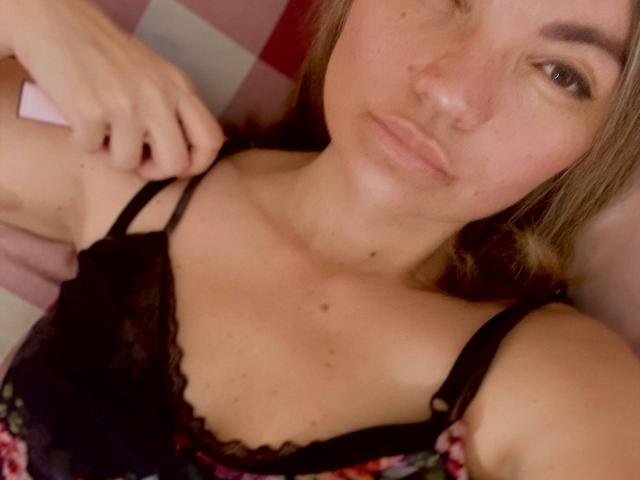 ElizavetaCream with pouty lips, wearing a black lace bra, has her hand on her shoulder, showing her small tits and freckles, with light brown hair and a relaxed expression.