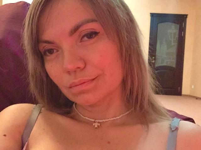 ElizavetaCream with light brown hair, a nose ring, and a subtle smile, wearing a blue strap dress, necklace with a small pendant, and showing her tits.