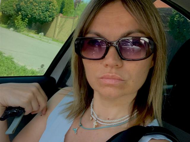 ElizavetaCream with brown hair, wearing black sunglasses, white tank top, and bead necklace, sitting in car, hand on steering wheel, serious face, seatbelt visible, car window in background.