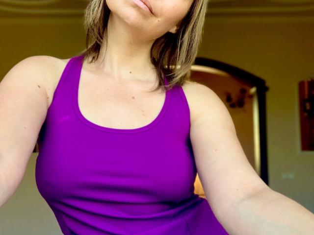 ElizavetaCream with medium boobs, wearing a purple tank top, her face slightly tilted, blonde hair, fair skin, and a subtle smile, in a bright indoor setting.