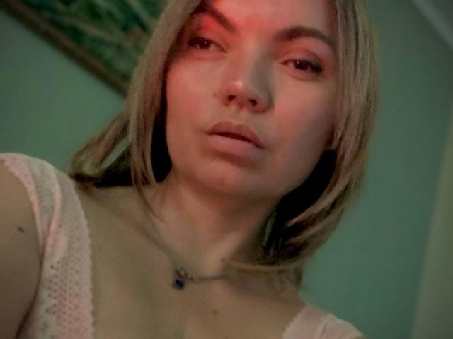 ElizavetaCream has blonde hair, pale skin, and is looking at the camera with a slight pout. She's wearing a white lace top, and has a small necklace.