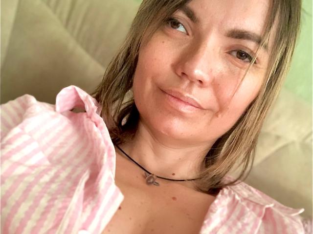 ElizavetaCream has a cute smile, brown eyes, and a slight tan with freckles. She's wearing a pink striped shirt and a black necklace with a small pendant.