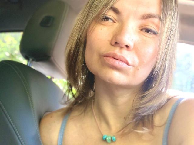 ElizavetaCream has light brown hair, pouty lips, and a blue necklace with a turquoise pendant. She's wearing a blue top, sitting in a car with green seats.