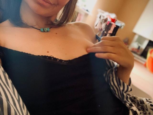 ElizavetaCream with bare shoulders, black lace-trimmed top, striped shirt off, holding phone, blue necklace, showing her tits, brown hair, slight smile, in a cozy room.