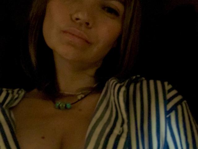 ElizavetaCream with a nose piercing, brown hair, and a striped shirt, showing cleavage with small moles, wearing a green bead necklace, in dim light.