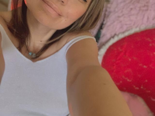 ElizavetaCream with light skin, brown hair, and a white tank top, smiling slightly, showing a small pimple on her cheek, taking a selfie, with a red pillow in the background.