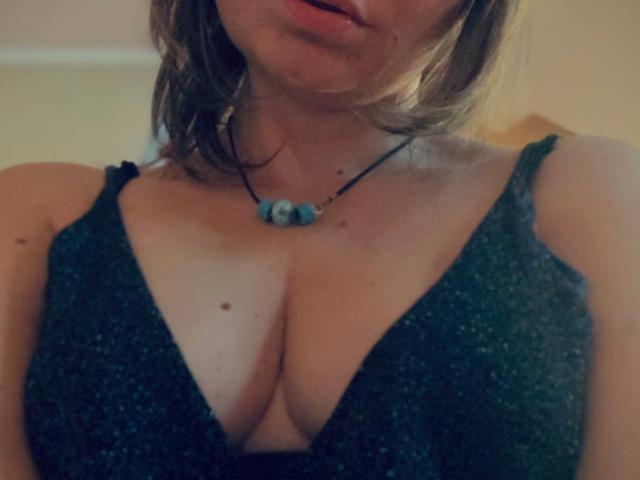 ElizavetaCream with medium tits in a deep green, sparkly dress, wearing a black necklace with a blue bead, showing cleavage and a small mole on her chest.