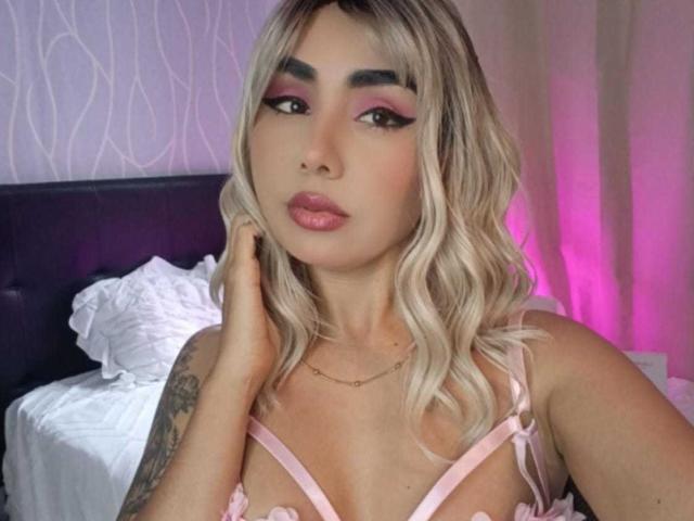 EmilyValk met blond, golvend haar, roze oogschaduw en roze lippenstift, draagt een roze beha, raakt haar gezicht aan en laat een tatoeage op haar rechterarm zien, in een slaapkamer..