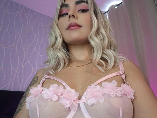 EmilyValk with blonde hair, pink eyeliner, and pink floral lingerie, showing her tits, tattoo on her right arm, and a slight smirk. Soft pink and purple lighting.