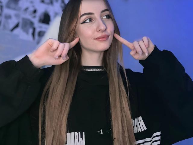 CutiCrystall-hot with long brown hair, black shirt, pointing at her pink cheeks with both fingers, blue-lit background, smiling slightly.