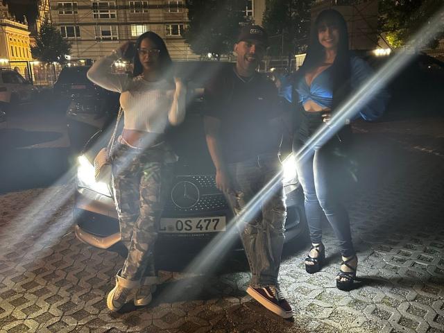 Three people stand in front of a car at night. TrioTemptation on the left wears a white crop top and camo pants, with a hand on her hip. The middle TrioTemptation wears a black cap, dark shirt, and camo pants, smiling. The right TrioTemptation wears a blue top, black pants, and strappy sandals, with a hand on her hip. Bright light flares from the car's headlights. The car has a license plate reading "OS 672." The trio is on a cobblestone street with buildings and trees in the background.