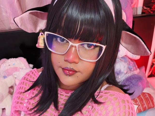 MissCurvyTop com cabelo preto, orelhas de gato, óculos rosa, piercing rosa no nariz, blusa de rede rosa e batom rosa, olhando para a câmera com os lábios rosa levemente entreabertos..
