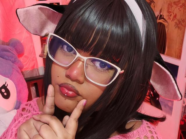 MissCurvyTop con cabello negro, orejas de gato blancas, gafas rosas y labios rosados brillantes, tocándose la barbilla, vistiendo una blusa de encaje rosa y una diadema blanca, en un selfie..