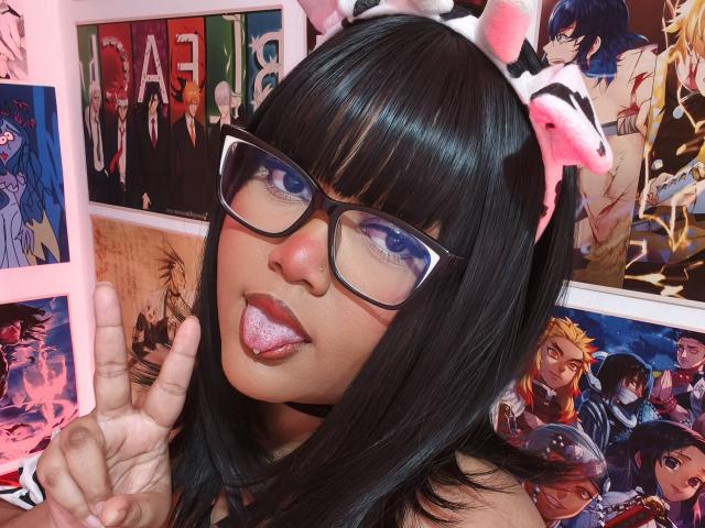 MissCurvyTop com cabelo preto, óculos, orelhas de gato rosa, língua de fora e piercing na língua, fazendo um sinal de paz. Pôsteres de anime ao fundo. Seios não visíveis..