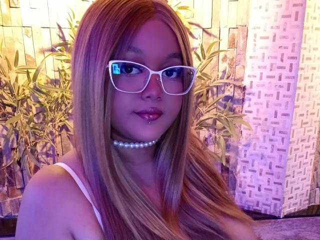 MissCurvyTop is a young woman with long, straight, blonde hair and fair skin. She wears white, thick-rimmed glasses and a pearl choker. Her lips are slightly parted with a small piercing on her lower lip. She is dressed in a white spaghetti-strap top. The image is lit with purple and yellow light, emphasizing her hair and glasses. Her expression is neutral, slightly looking to the side. The background has a bamboo pattern with a textured wall.