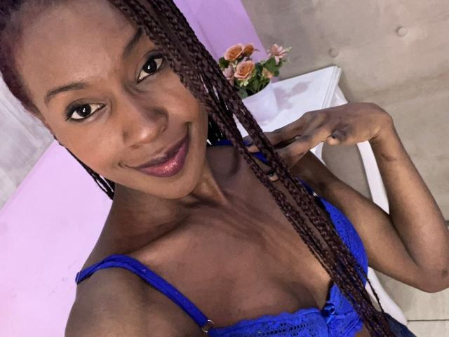 AliisLove, de pele escura, com tranças longas, sorrindo, usando um sutiã de renda azul, mostrando seios pequenos, segurando uma trança, batom rosa, parede roxa ao fundo..