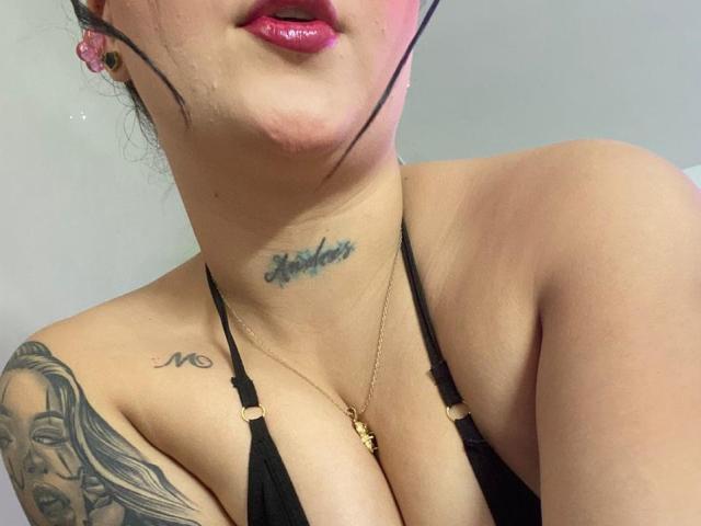 MelodyBlossom with pink lips, black halter top, tattoo under chin, tattoo of woman's face on shoulder, necklace, black hair strands, light skin, visible cleavage.
