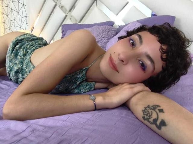 EmilyAllen with curly brown hair, purple eyeshadow, green patterned top, lying on purple bed, tattoo on right forearm, bracelet on wrist, looking at camera, relaxed expression.
