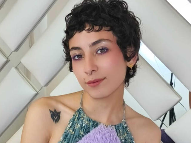EmilyAllen has short curly black hair, purple eyeshadow, a nose ring, and a small black tattoo on her left shoulder. She's wearing a colorful halter top.
