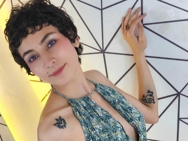 Emily Allen, avec ses courts cheveux noirs bouclés, son fard à paupières violet, son anneau au nez et ses tatouages sur les deux épaules, porte un haut à bretelles bleu à motifs et sourit à l'appareil photo..