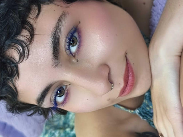 EmilyAllen's face, curly black hair, blue eyeliner, nose ring, small lip piercing, light skin, subtle blush, gentle smile, looking up, hand resting on cheek, pink lips, green eyes.