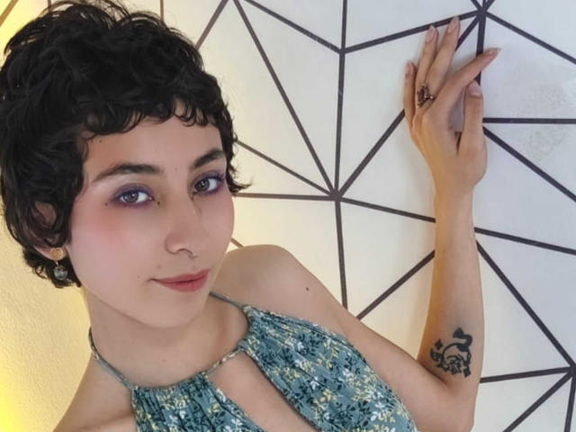 Emily Allen, avec ses courts cheveux noirs bouclés, son haut à encolure plongeante floral bleu-vert, un anneau au nez, du fard à paupières violet, un tatouage sur l'avant-bras droit, et les mains contre un mur géométrique, souriant légèrement..