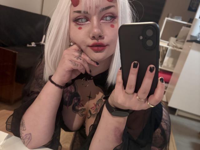 PerfectGabbie takes a selfie with a black smartphone. She has white hair with red horns, pink eyeshadow, red lipstick, and red facial marks. Her black nails are short and polished. She has a septum ring, a lip ring, and wears a black choker with a ring. Her right hand has tattoos and rings, and she has a tattoo on her left arm. She has a black sheer robe, and a tattoo on her chest. Her phone case has a large camera hole. The photo has a casual, intimate feel.
