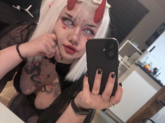 PerfectGabbie takes a selfie with a black smartphone. She has white hair, red devil horns, and black eye makeup with red heart-shaped marks on her cheeks. She's wearing a black sheer robe, exposing tattoos on her left arm and chest. Her nails are black-painted, and she has a nose ring. Her left hand touches her cheek. The background is a blurred kitchen. Her phone case has a black camera lens cover. She's wearing a black bracelet and a gold ring. The photo is candid and focused on her face.