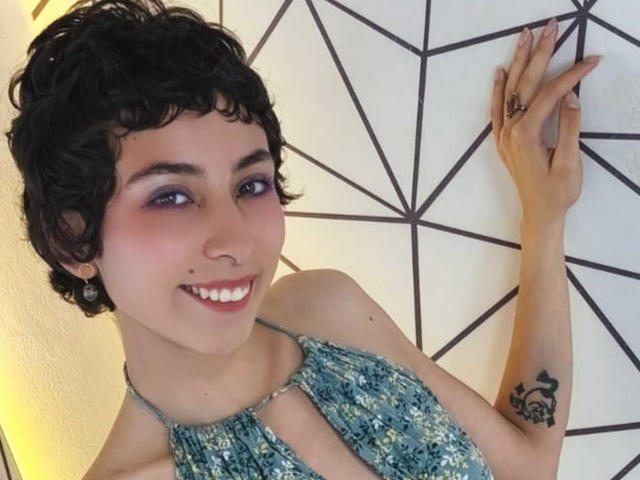Emily Allen, met kort krullend zwart haar, paarse oogschaduw, lachend, draagt een blauw gestreept topje met halter, toont een tatoeage op haar rechterpols, met haar hand tegen een geometrische muur..