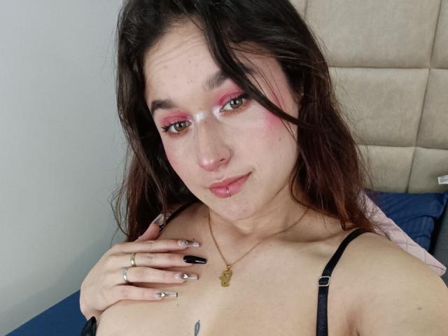 MilaGonz with pink eyeshadow, black nails, and a gold necklace, touching her tits, has a small tattoo on her chest, and a slight smile. Cum on her face.