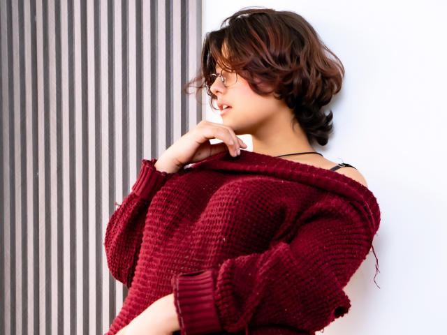 MisakiWong with wavy brown hair, glasses, and a red knit sweater, hand near mouth, showing her tits, against a white wall with striped black-and-white background.