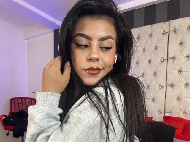 CamilePretty with long black hair, thick eyebrows, and red lipstick, wearing a gray hoodie, holding her hair, and wearing white earbuds, looking down.