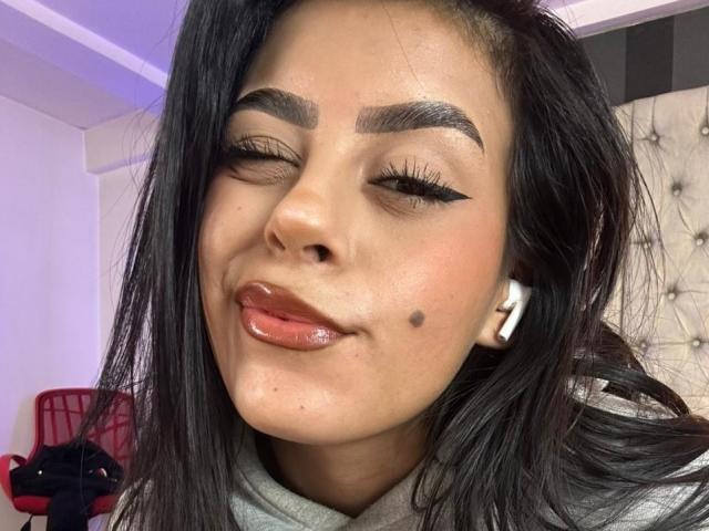 CamilePretty has shiny black hair, thick eyebrows, and a small beauty mark on her right cheek. She's winking with glossy lips and wearing white earbuds.