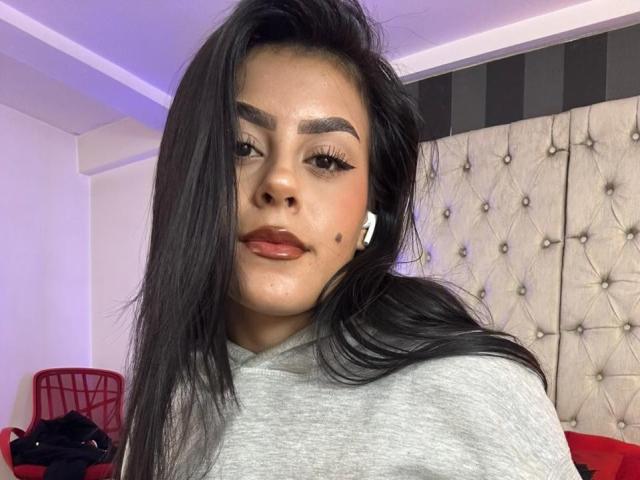 Close-up photo of CamilePretty with long, straight black hair, brown skin, and dark eyebrows. She wears a gray shirt, has red lipstick, and a small beauty mark on her right cheek. She has white earbuds in her ears. The background includes a tufted beige headboard, a red chair, and a black and white striped section on the wall. The lighting is soft with a purple-blue tint. The composition focuses on CamilePretty's face and upper shoulders.