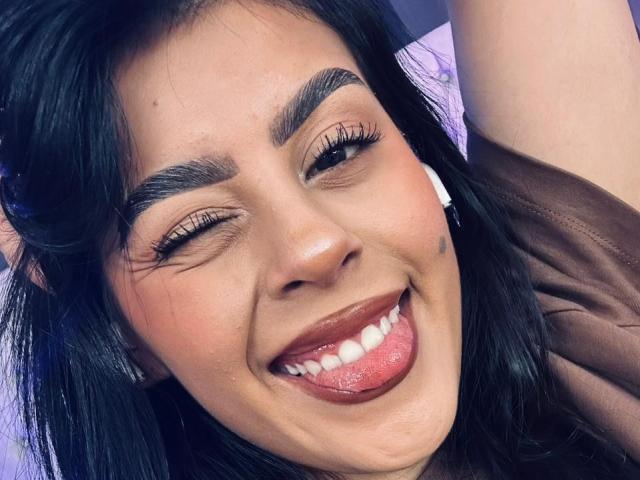 Close-up of CamilePretty with dark, wavy hair, brown eyes, and thick, well-groomed eyebrows. She's winking and sticking out her tongue, revealing white teeth and a pink tongue. She has long, dark eyelashes and wears subtle makeup with a light pink blush. Her skin is light brown and she's wearing a brown garment. The image has a slightly blurred, purple background. Her expression is playful and confident.