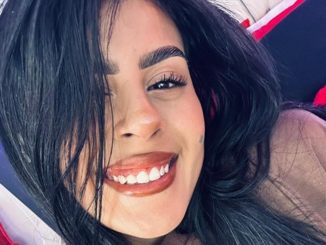 Close-up photo of CamilePretty with dark, wavy hair partially covering her face. She has thick, arched eyebrows, long, dark eyelashes, and is smiling with visible white teeth and glossy, dark red lipstick. Her skin is light brown, and the image focuses on her smiling face. The background is blurred, with red and white elements. The texture of her hair is smooth and slightly shiny. Her expression is confident and cheerful.