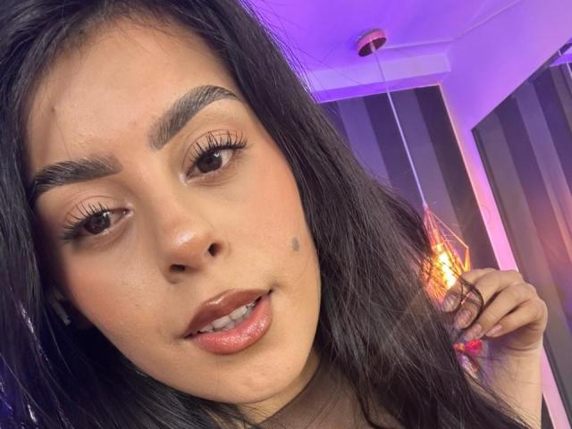 CamilePretty has dark eyebrows, long black hair, and a small beauty mark on her left cheek. Her glossy pink lips are slightly parted. She's looking to the side.
