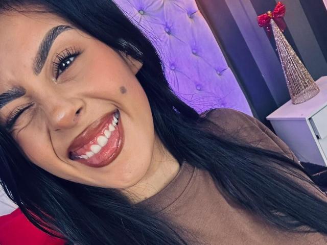 CamilePretty, with black hair, winks and sticks out her tongue, showing white teeth and a small beauty mark on her left cheek. She wears a brown top and a nose ring.